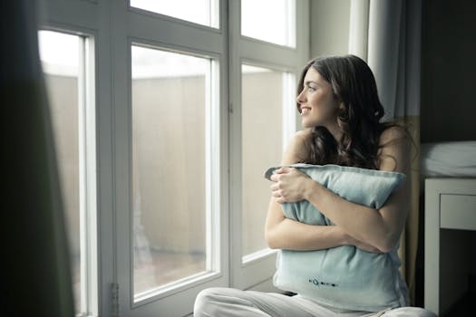 pexels-photo-920386-920386 A woman sits by the window, hugging a pillow while smiling warmly.
